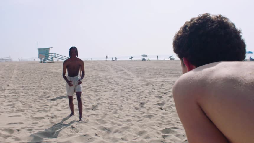 On a wide stretch of sand a shirtless man in pale board shorts walks toward the camera while another persons sanddusted back fills the foreground with a pastel lifeguard tower distant houses and scattered beachgoers