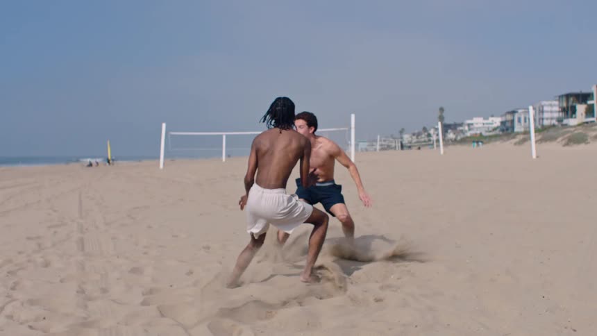 Young man runs across the beach carrying a football as another young man fails to tackle him