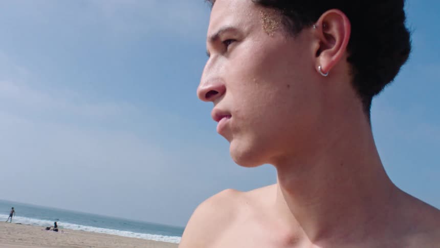 A young man holds and grips a football on a beach with other people in the background