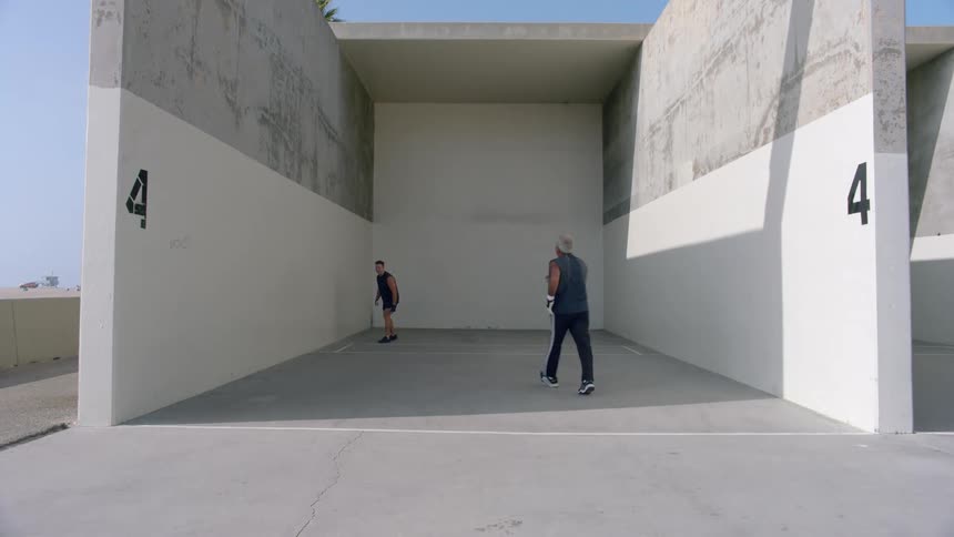 On a sunny day an older man and a younger man play a competitive game of handball running and hitting the ball against the concrete walls of the outdoor court