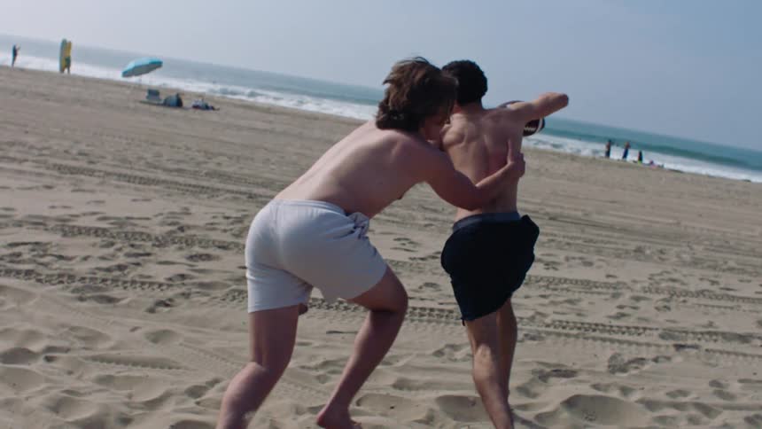 During an energetic game of beach football one friend tries to dodge the other but is tackled and they both tumble into the soft sand by the ocean