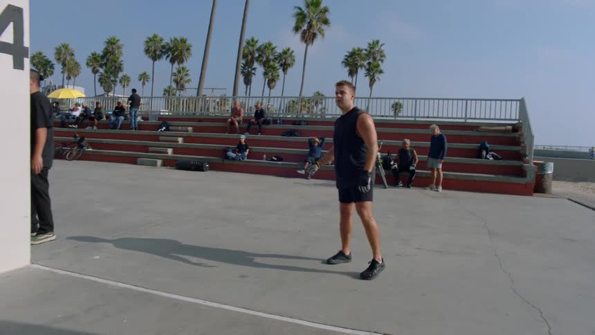 A man in athletic gear stands on a concrete outdoor court and performs an underhand serve against a numbered wall while several people watch from the nearby bleachers under a clear sky