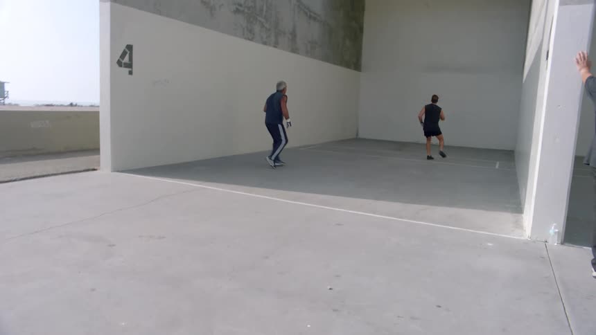Two men throwing a small ball against a white wall in an outdoor space