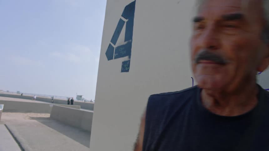 A senior man with a mustache and grey hair stands in profile leaning against a white concrete wall marked with a blue number four and a blue sky in the background