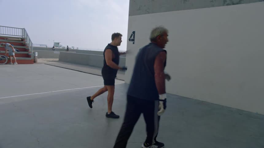 Two men wearing gloves are throwing a small blue ball against a white wall in an outdoor space