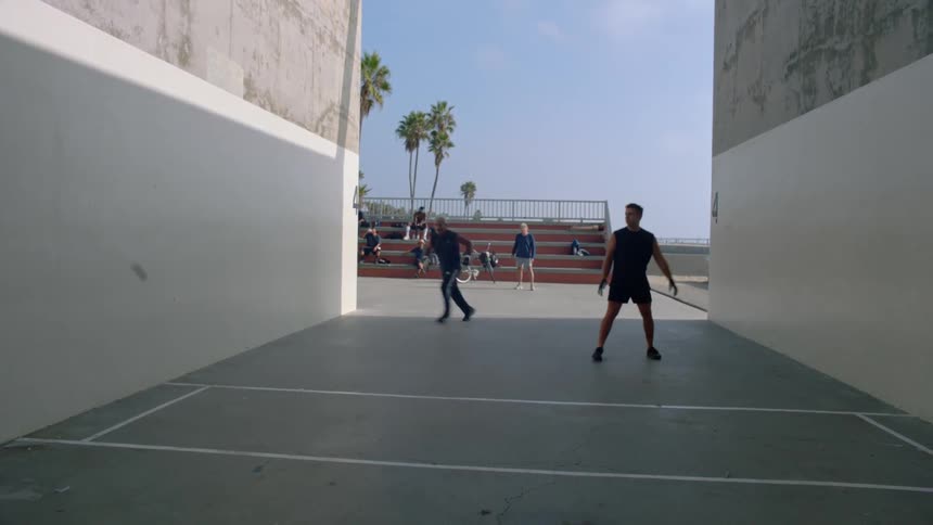 From inside an openair court two men play a spirited game of handball while spectators watch from nearby bleachers under tall palm trees