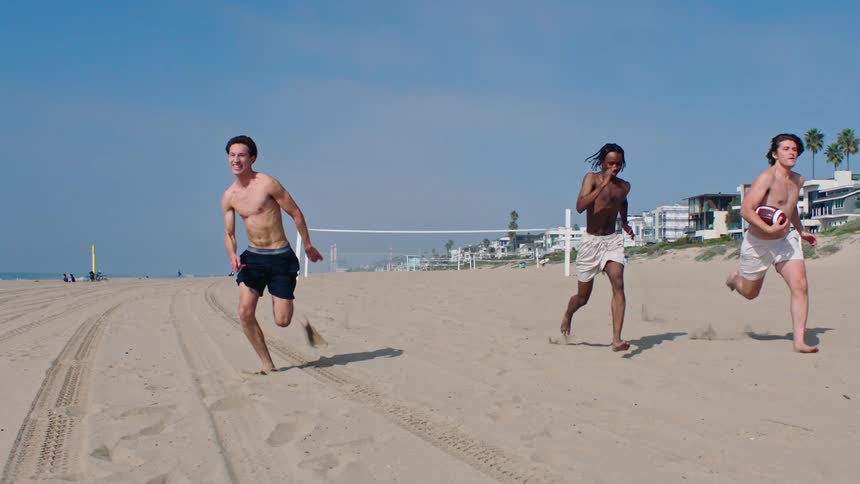 Three men running on the beach sand one of them holding an american football