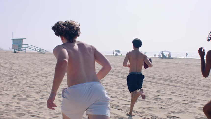 Three boys run across the beach while playing with a football on a summer day