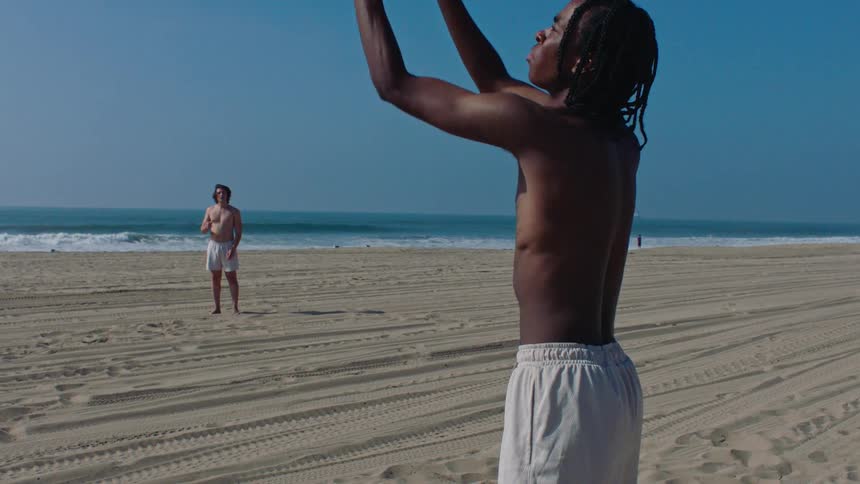 A black man catches a football on a sandy beach while another man stands in the background