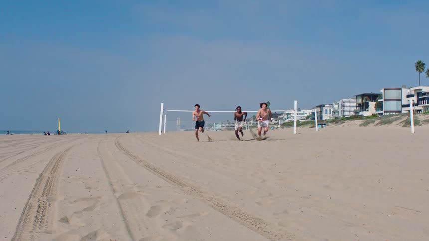 Three teenage boys racing shirtless across the beach sand on a sunny day
