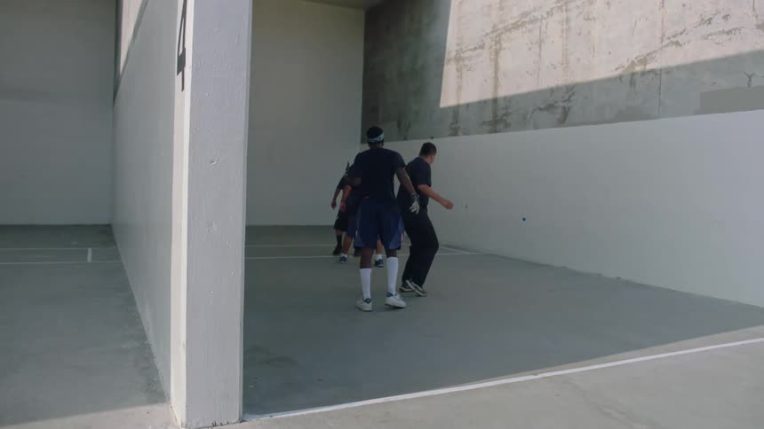 A group of men is seen playing a game of handball on an outdoor concrete court under bright sunlight one player hits a small blue ball against the high wall while the others move into position to continue the match