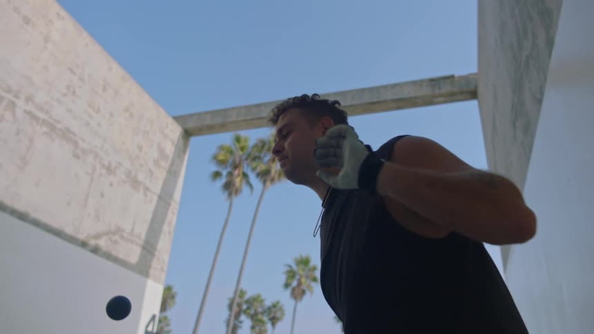 A man wearing padded gloves serves a handball with an underhand motion against a tall concrete wall while standing in an outdoor court framed by palm trees and a clear blue sky