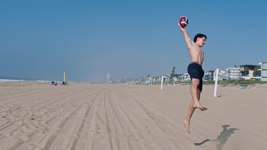 A shirtless young man runs across a wide sandy beach and leaps high into the air to catch a football before falling onto the sand