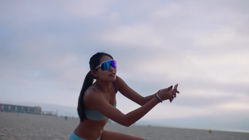 On a cloudy beach a focused volleyball player in athletic swimwear and reflective sunglasses lunges forward with outstretched arms to bump a yellow and black ball midair