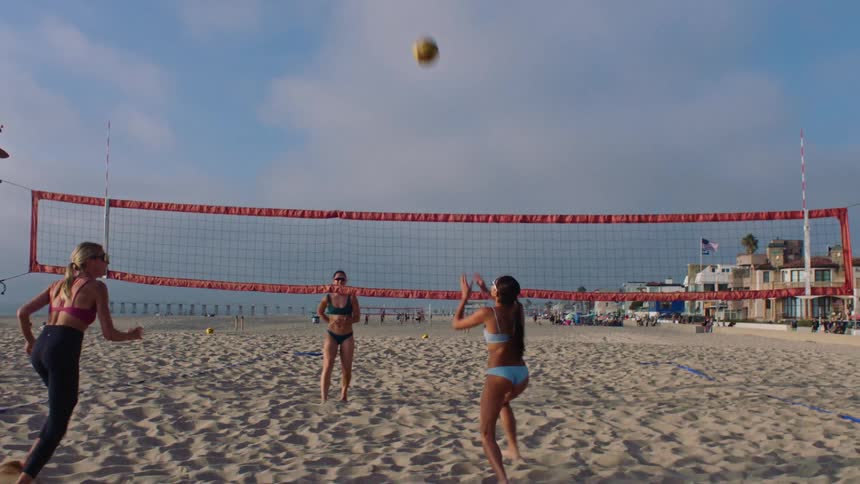 Group of four women playing volleyball in pairs on beach sand at midday all wearing swimsuits