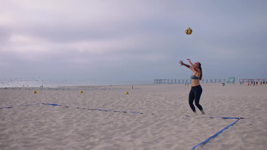Woman wearing sunglasses throws a volleyball onto a sand court at the beach