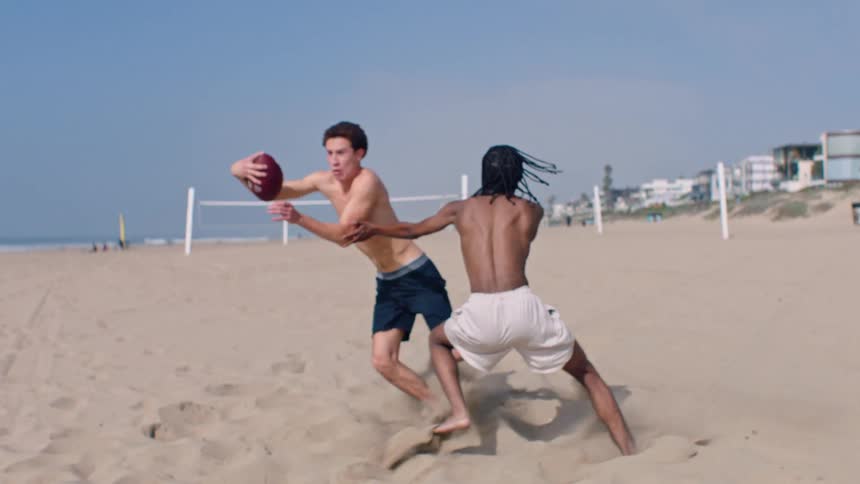 Two shirtless young boys play football on a wide sandy beach with one player carrying the ball and performing a quick juke move to successfully evade a defender who falls while trying to make a tag