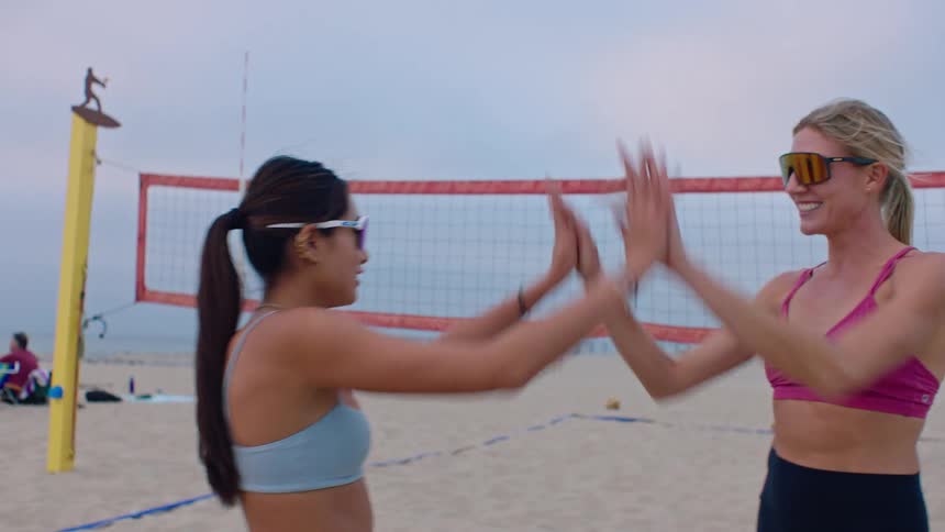 Two women wearing sports bras and sunglasses celebrate on a sand court by giving each other a highfive in front of a volleyball net with the ocean and a pier visible in the hazy background