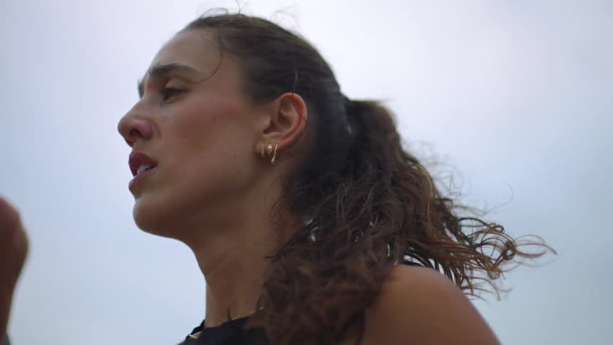 A close view from below shows a woman with dark curly hair tied in a ponytail looking upward with a serious expression against a pale sky