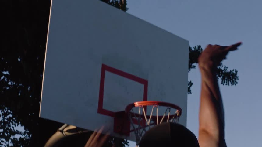A man wearing a black cap jumps up and successfully dunks a basketball into a hoop on an outdoor court the camera captures the action from behind the player showing the ball passing through the net against a clear sky