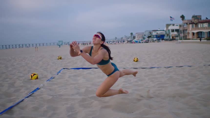 A woman makes a dynamic play in beach volleyball running and diving into the sand to bump the ball before looking up with a smile
