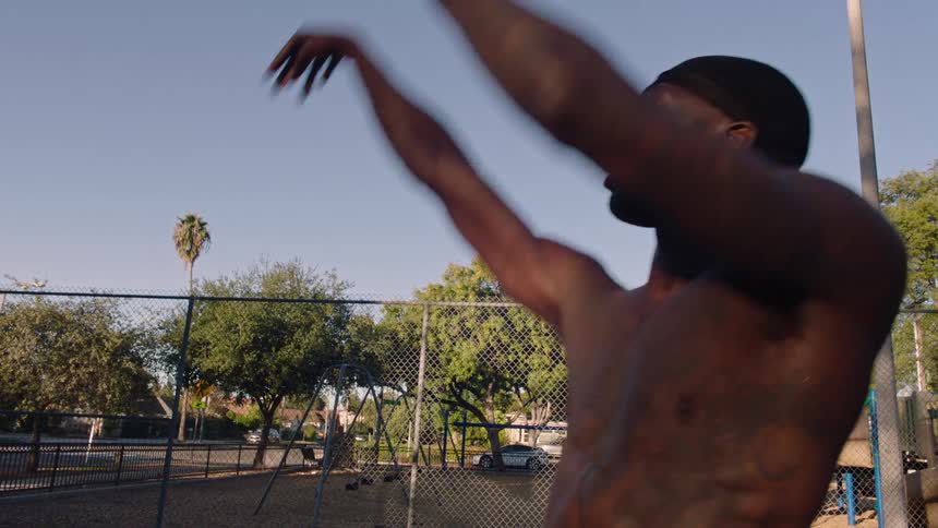 A shirtless man wearing a black durag shoots a basketball on an outdoor court surrounded by a chain link fence and green trees under a clear blue sky