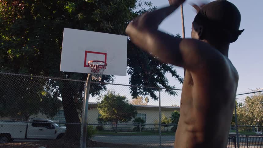 A shirtless man wearing a black durag stands on a blue outdoor basketball court and shoots a ball into a hoop during a clear day