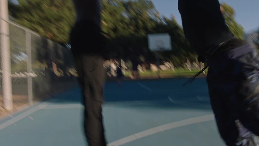 On a sunny treelined park court a shirtless player drives the ball toward the baseline while a defender in a black tank and joggers slides to contest long shadows streaking across the painted lines with distant mountains in view