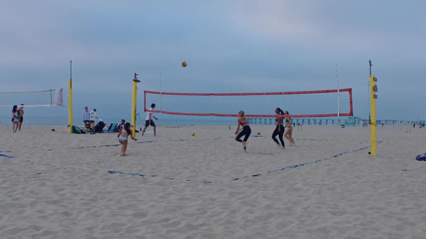 Several women play beach volleyball on a sandy court near the ocean with many beachgoers with two players at the net and others positioned behind them under an overcast sky