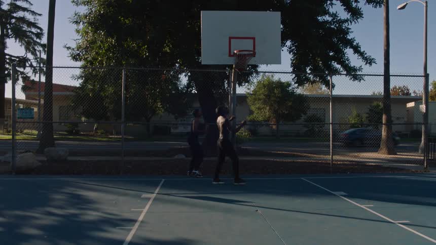 Two men play a competitive oneonone basketball game on a blue outdoor court as one player dribbles past a defender to score a layup under the afternoon sun