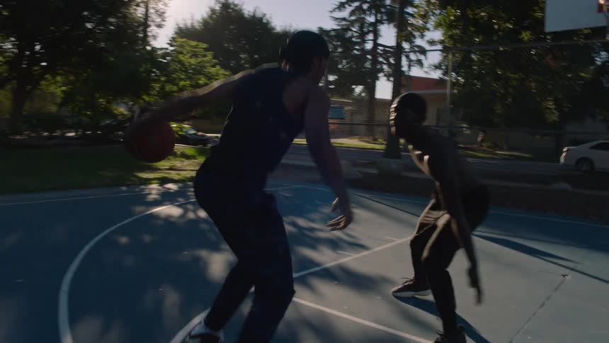 Two men play basketball on an outdoor court during sunset and one player dribbles past a defender and executes a jump shot toward the hoop while lens flares streak across the frame