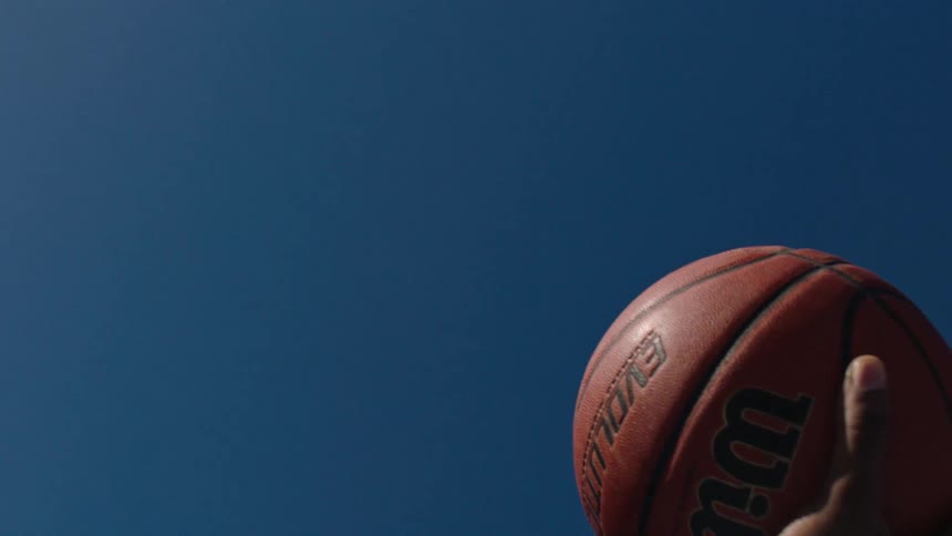 A persons hand throwing an orange basketball upwards contrasts with the blue sky