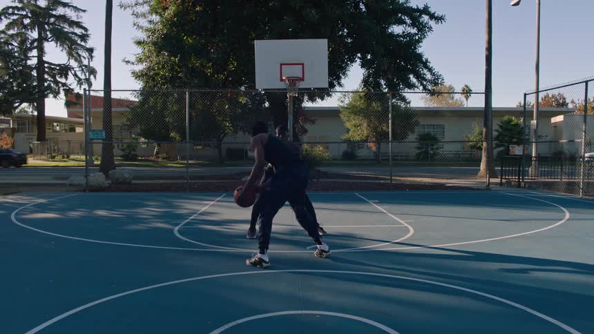 In this video two men play a game of oneonone basketball on an outdoor blue court during the daytime one player successfully drives past his opponent to the hoop while the other watches the shot from the perimeter