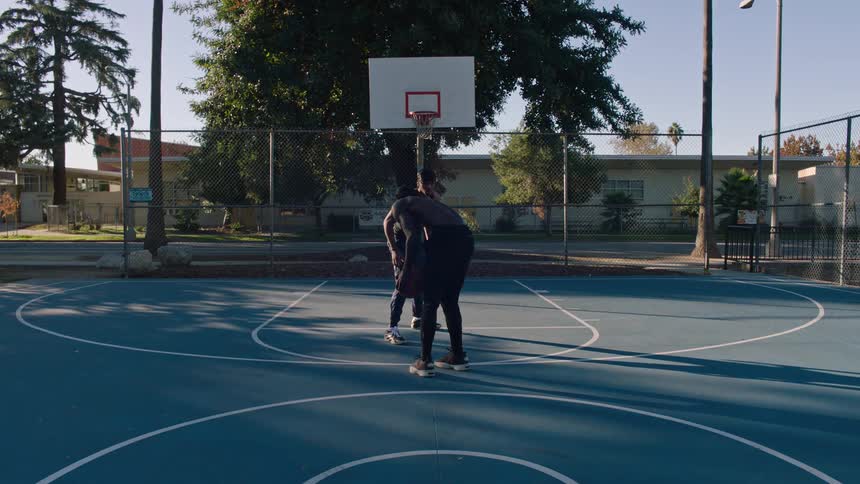 Two men play basketball on an outdoor court with one shirtless player jumping to shoot the ball while the other defends beneath the hoop