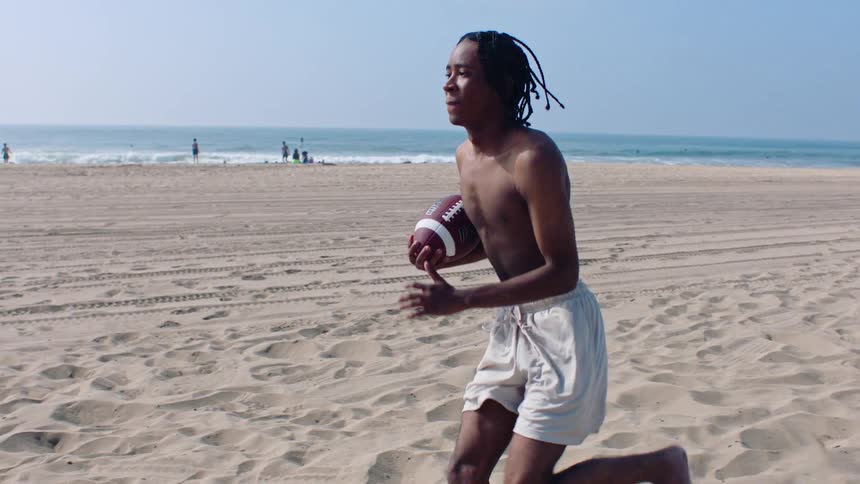 A man is holding an american football running on the sand of a beach when another man tries to intercept him