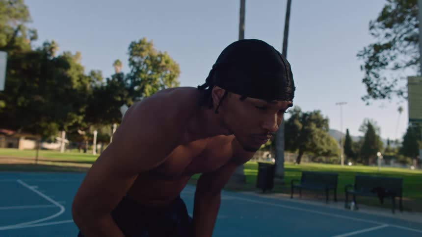 A young shirtless man wearing a black beanie is holding a basketball staring straight ahead in a sporty stance