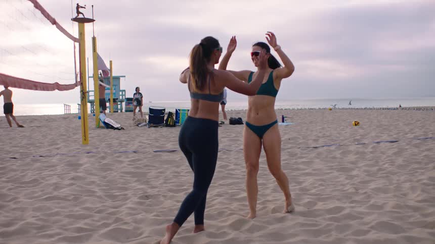 On a cloudy beach two women in athletic swimwear and sunglasses highfive between points on a sand volleyball court beside yellow poles and a net with a lifeguard tower scattered gear other players and the ocean in the background