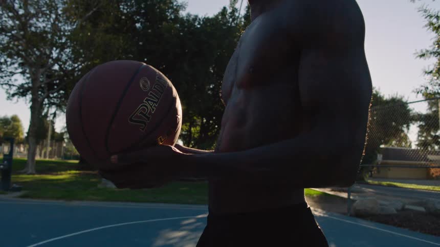 A man holds a basketball while standing outdoors on a court during golden hour