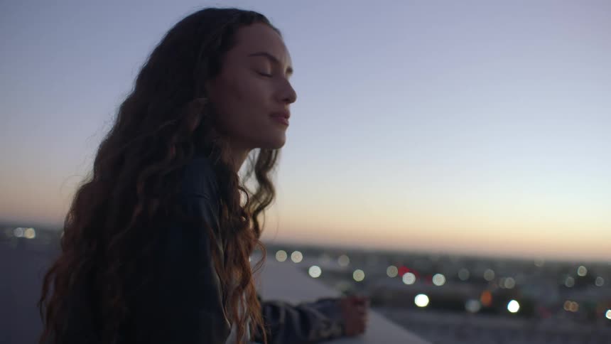 A young woman with long curly brown hair stands on a rooftop at dusk she looks out over a city skyline as the sun sets her hair blowing gently in the breeze