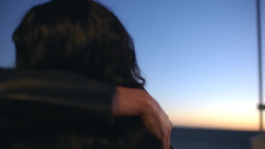 Wo women in dark jackets share a long supportive hug on an outdoor rooftop as the sun sets showing calm and affectionate expressions against a blue twilight sky