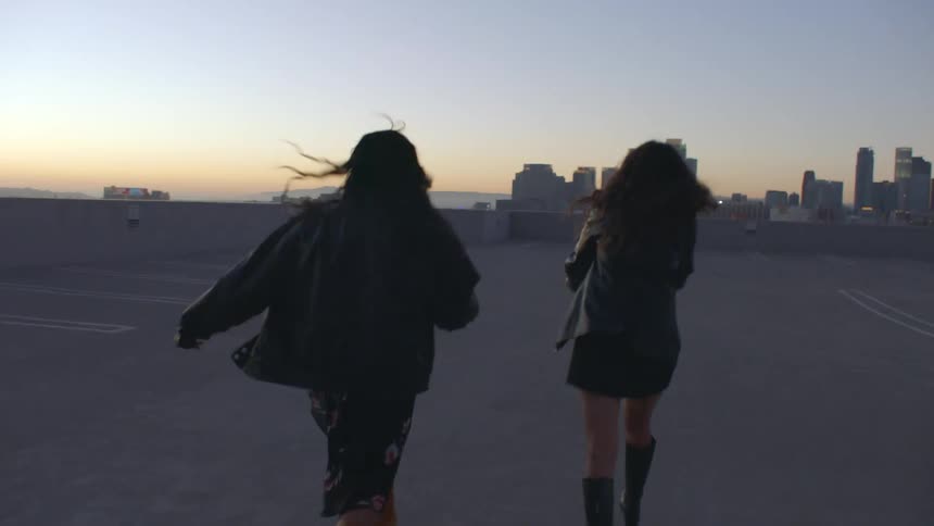 Two young women in leather jackets and boots walk across an empty rooftop parking lot at dusk spinning and laughing with a sprawling city skyline visible in the distance during the golden hour