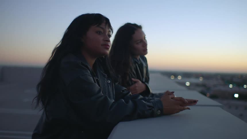 Two women in leather jackets lean against a concrete ledge on a rooftop at dusk looking out over a sprawling city as the sun sets on the horizon