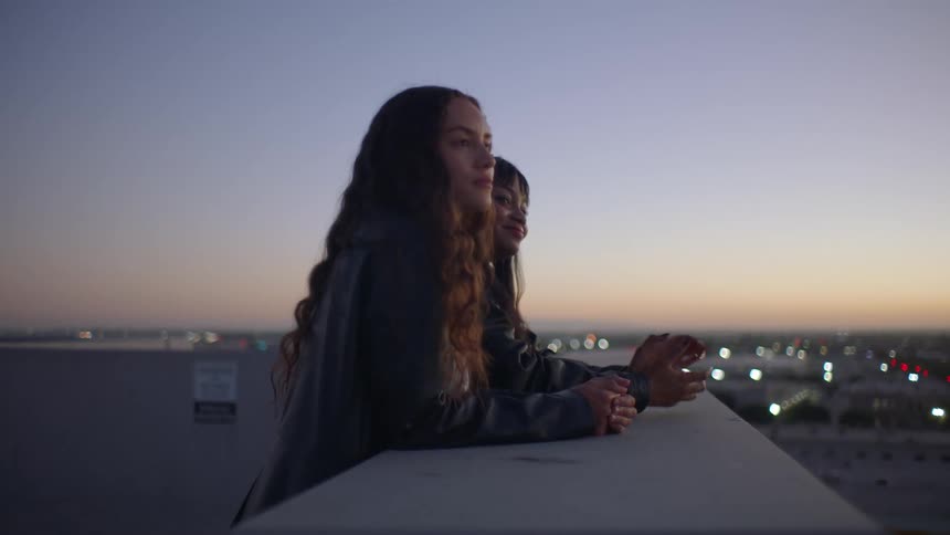 Two women lean against a concrete ledge on a rooftop at twilight looking out over a blurred city landscape as the sky glows with the last remnants of sunset