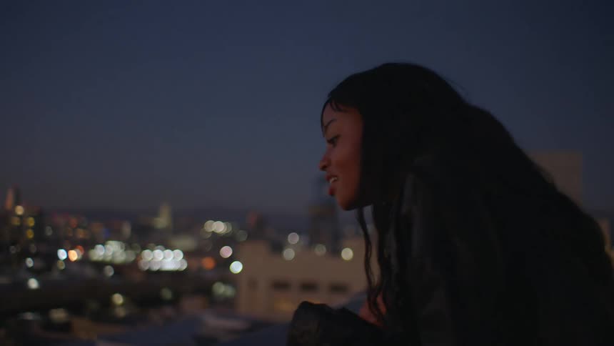 Young woman with braided hair standing on a rooftop at night smiling as she looks down at the busy street below
