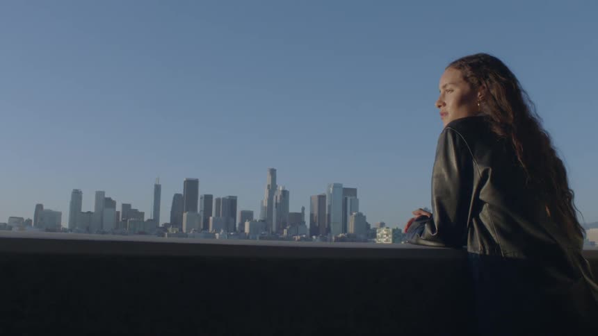 A woman with long flowing hair leans on a ledge looking out over a sprawling city skyline that fills the horizon under a clear sky