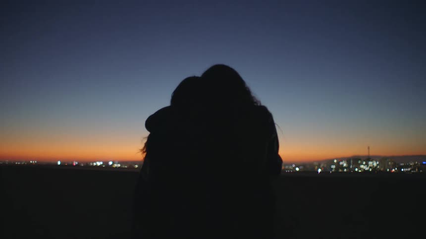 Two silhouetted friends embrace overlooking a glowing city skyline at dusk