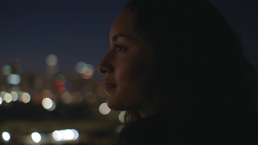 Young woman standing outdoors at night smiling briefly as she looks curiously to her left with blurred city lights behind her