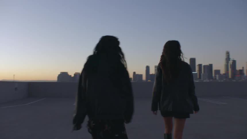 Two young women walking side by side in black jackets at sunset with the city skyline behind them