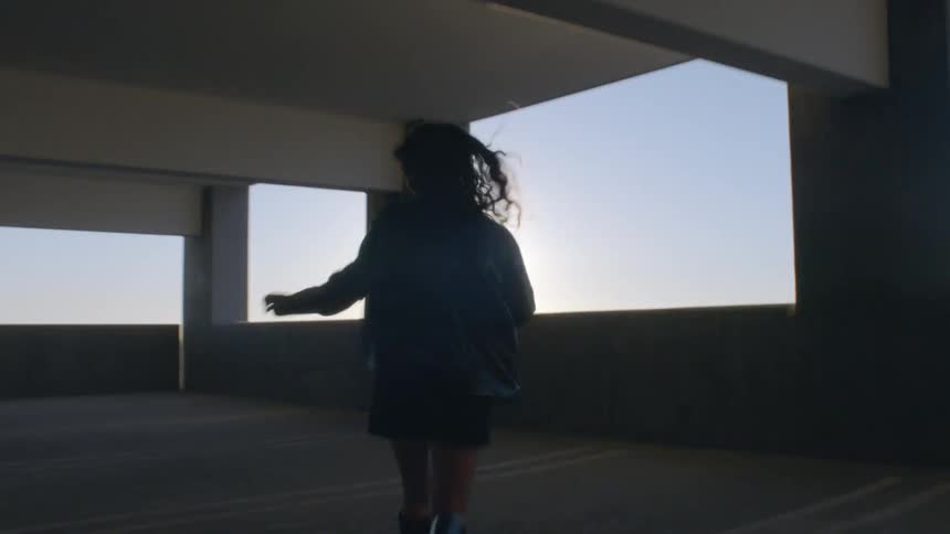 A woman with long hair runs through a dark parking garage toward a bright sunset her silhouette framed by the golden light before she reaches the edge to look out at the city skyline