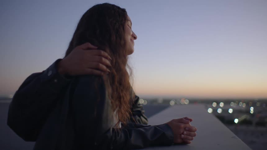 Two young women standing on a rooftop at sunset one smiling as she puts her arm around the others shoulder